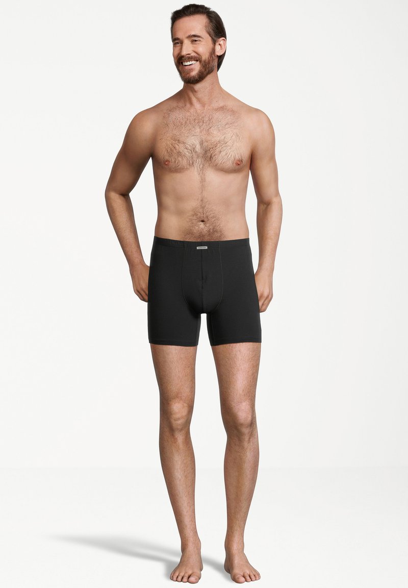 Boxer brevi neri con un design aderente e una fascia elastica in vita, indossati da un uomo in piedi a piedi nudi su uno sfondo neutro.