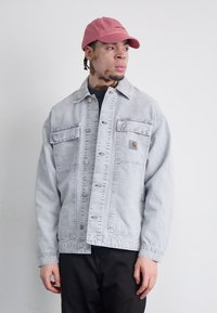 Light grey denim jacket with button front, two chest pockets, and logo patch; paired with a pink cap and black pants. Smooth texture.