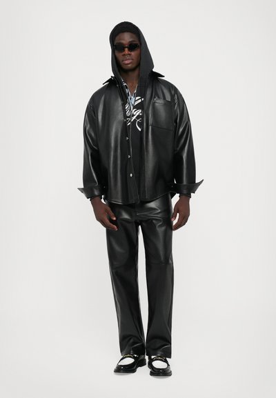 Homme portant une veste en cuir noir et un pantalon noir avec un sweat à capuche noir et des lunettes de soleil, se tenant contre un fond blanc uni.