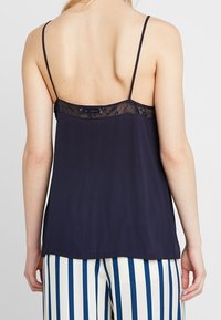 Marineblaue Camisole mit dünnen Trägern, die ein Spitzenpaneel am oberen Rückenbereich aufweist. Der Stoff ist glatt und leicht, kombiniert mit gestreiften Hosen.
