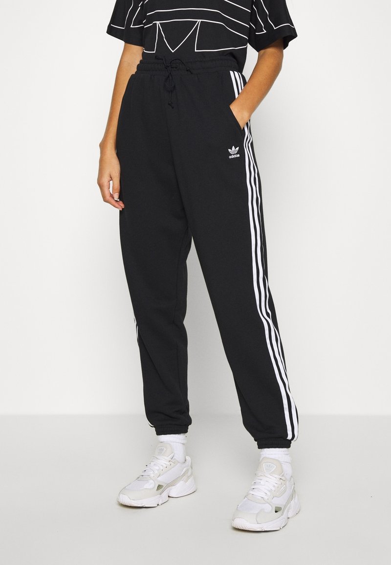 Pantaloni della tuta neri con strisce laterali bianche, vita elastica e tasche, abbinati a sneaker bianche. Materiale morbido e vestibilità rilassata.