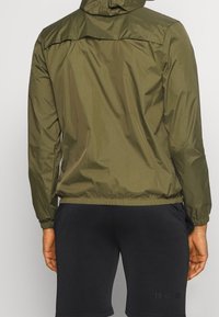 Giacca leggera verde oliva con cappuccio, polsini e orlo elastici; indossata sopra pantaloni corti neri con un sottile dettaglio grafico sulla gamba.
