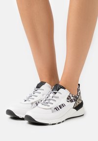 Weiße Sneakers mit schwarzen Sohlen, Details im Leoparden- und Zebramuster, schwarz-weiße Schnürsenkel, die auf nackten Füßen vor einem schlichten Hintergrund getragen werden.
