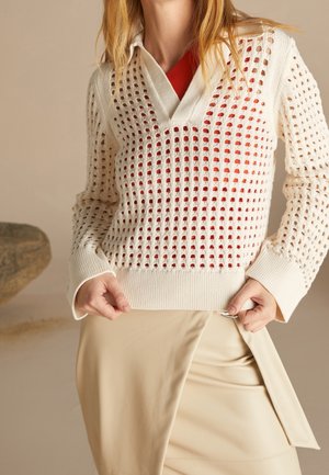 Femme portant un pull en maille ouverte crème sur un haut rouge, assorti à une jupe portefeuille beige, mains tenant l'ourlet du pull.