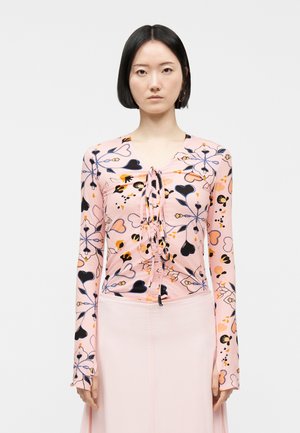 Vrouw met steil zwart haar, gekleed in een roze top met lange mouwen met blauwe en oranje bloemenpatronen en een bijpassende lichtroze rok.