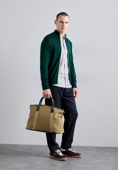 Cardigan vert, chemise à fleurs, pantalon bleu marine, baskets marron. Portant un sac en toile beige avec des accents noirs et beiges.