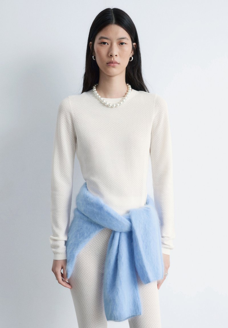 Haut à manches longues texturé crème associé à des leggings crème ajustés et un châle doux bleu clair. Collier de perles et boucles d'oreilles argentées en forme d'anneau.