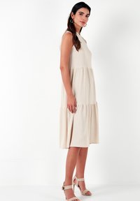 Beige maxi dress with a tiered design, sleeveless cut, and textured fabric. Paired with neutral heeled sandals.