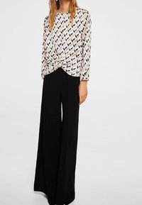 Langarm blouse met een geknoopte voorkant, versierd met een bladpatroon in roze en zwart op een witte achtergrond. Gecombineerd met wijde zwarte broeken.