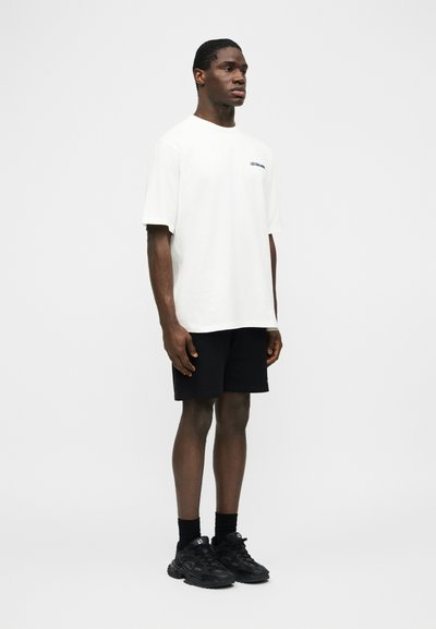 Mann steht und trägt ein weißes übergroßes T-Shirt, schwarze Shorts, schwarze Socken und schwarze Turnschuhe vor einem schlichten hellen Hintergrund.