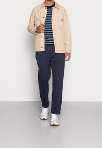 Beige bomullskavaj med bröstfickor, marinblårandig t-shirt, marinblå byxor och vita sneakers med bruna detaljer.