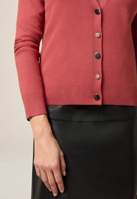 Valkoinen vadelmapinkki neulejakku, jossa on V-pääntie, kuusi nappia ja ribattu hihansuua. Se on puettu mustan nahkahameen päällä, joka on sileätekoinen ja tiukkahko muotoilu.