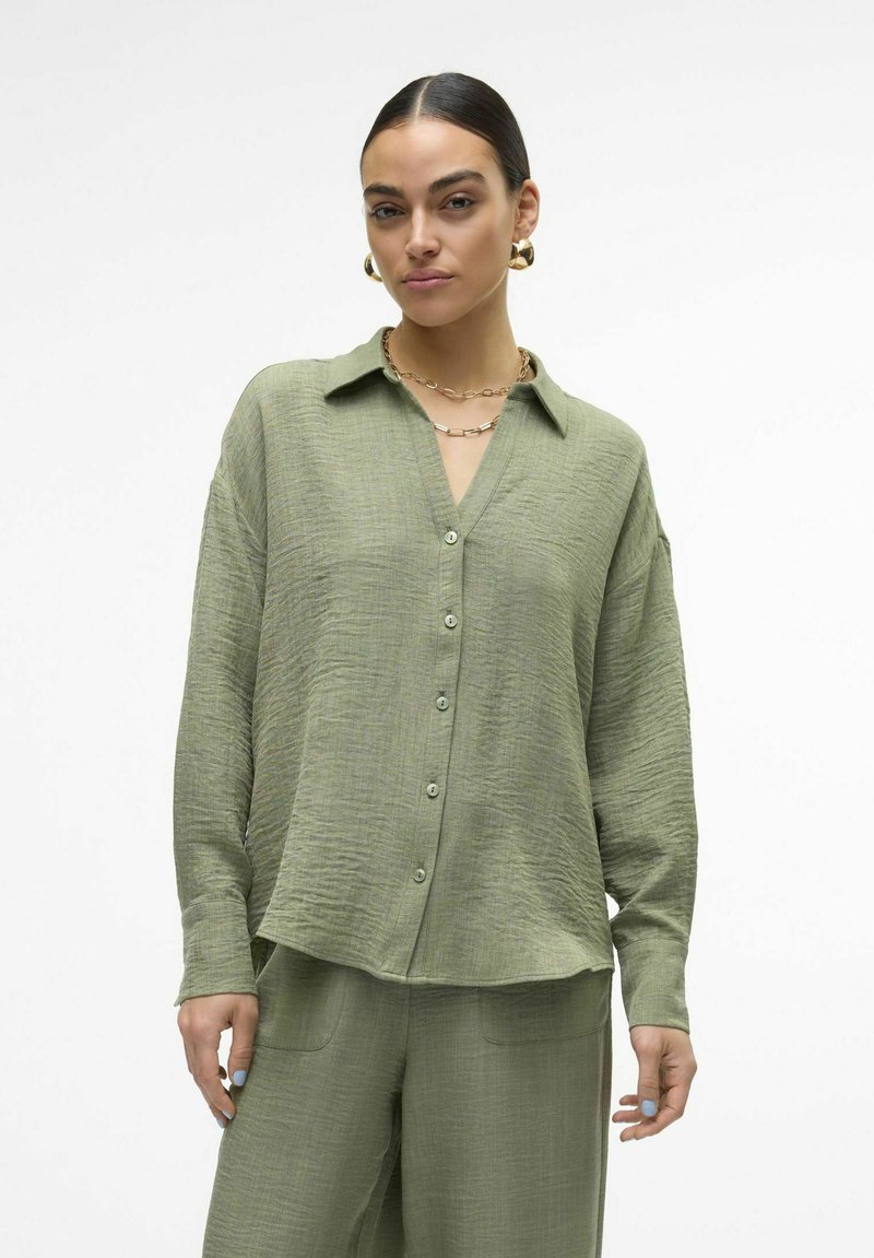 Camicia button-up verde chiaro, strutturata, con colletto, caratterizzata da un design a scollo a V e una vestibilità comoda. Il materiale sembra traspirante e morbido.