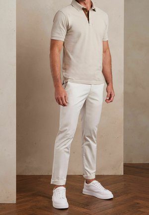 Beige polo shirt with a brown collar, paired with white tapered pants and white sneakers. Smooth fabric and fitted design with short sleeves.