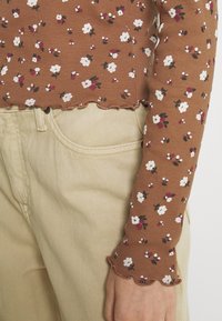 Braunes, geripptes Oberteil mit weißem und burgunderfarbenem Blumenmuster; mit gewelltem Saum und langen Ärmeln. Kombiniert mit beigen Hosen.