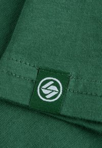 Groene katoenen stof met een gladde textuur. Heeft een gestikte zoom en een label met een wit logo op een groene achtergrond.