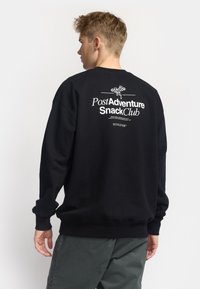 Sudadera negra con texto gráfico blanco "Post Adventure Snack Club" y un elemento de diseño en la parte posterior. Ajuste holgado con puños de canalé.