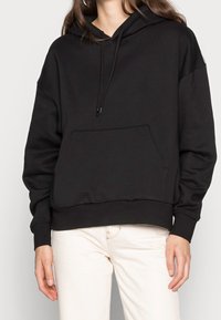 Mujer con una sudadera negra con bolsillo frontal y cordones, combinada con pantalones blancos frente a un fondo liso.