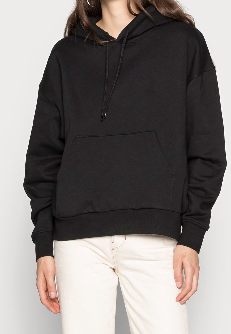Mujer con una sudadera negra con bolsillo frontal y cordones, combinada con pantalones blancos frente a un fondo liso.