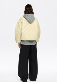 Gevoerde gele gewatteerde jas met een grijze capuchon, laag over een grijze sweatshirt, gecombineerd met brede zwarte broek. Gladde afwerking en oversized pasvorm.