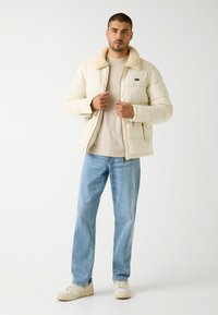 Giacca puffer beige con colletto in shearling, tasche con zip, indossata sopra una camicia chiara e jeans blu, completata da scarpe da ginnastica chiare.