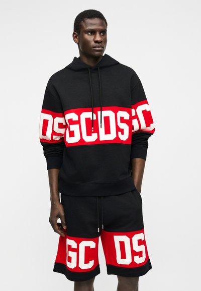 Uomo che indossa una felpa con cappuccio e pantaloncini neri con grandi scritte rosse e bianche GCDS su petto, maniche e gambe, in piedi con le mani nelle tasche.