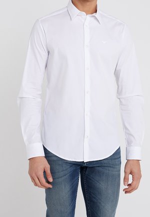 Homme portant une chemise blanche ajustée à manches longues avec col et un jean bleu, debout devant un fond uni.