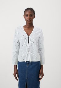 Λευκή δανδής μπλούζα με V-λαιμό, μακριά μανίκια και λεπτομέρεια με φιόγκο μπροστά. Διαθέτει λουλουδένια μοτίβα και μικρές μαύρες λεπτομέρειες.