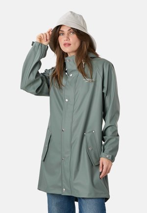Woman wearing a sage green raincoat with snap buttons and flap pockets, paired with a light beige bucket hat, touching the hat's brim.