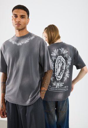 T-shirt cinza oversized feito de algodão, com bordado floral na frente e uma graphic da Virgem de Guadalupe nas costas.