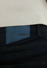 Tummat farkut, joissa on näkyvä sininen nahkatägi, jossa lukee "CHASIN'" vyötäröllä, ja teksturoitu beige neuloskangas yllä.