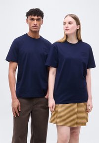 Άντρας και γυναίκα στέκονται δίπλα δίπλα φορώντας ταιριαστά μπλε ναυτικού χρώματος μπλουζάκια· ο άντρας φορά καφέ παντελόνι, η γυναίκα φούστα με κίτρινο καρό σχέδιο.