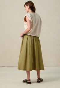 Haut sans manches beige clair avec de larges emmanchures, associé à une jupe longue et froncée de couleur vert olive. Porté avec des sandales marron foncé.