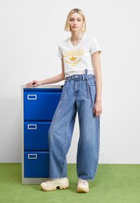 Balta trumpa T-shirt su geltonu Adidas logotipu, aukšta juosmens šviesiai mėlynos spalvos plačiakelnėmis džinsais ir kremine masyviais sportbačiais, šalia mėlynos dokumentų spintos.
