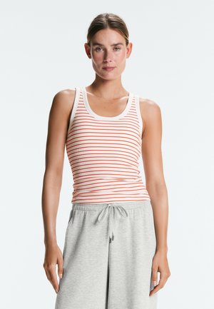 Žena v bílem tílku bez rukávů s tenkými oranžovými vodorovnými pruhy a světle šedých kalhotách se stahovací šňůrkou, stojící před neutrálním pozadím.