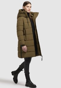 Abrigo acolchado verde oliva con capucha, diseño acolchado y cierre de cremallera. Combinado con pantalones negros ajustados y botas de tobillo.