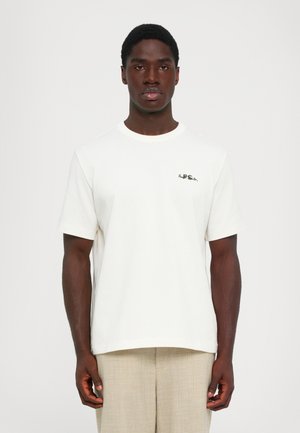 Sort mand iført en hvid T-shirt med korte ærmer og lille sort tekst på brystet samt beige bukser, der står foran en ensfarvet baggrund.