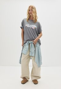 Ung kvinde med bølget blond hår iført en oversized grå T-shirt, beige vide bukser, en stribet skjorte bundet om livet og brune mokkasiner.