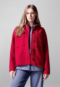 Röd texturerad jacka med två stora bröstfickor, svarta knappar och en krage, bärs över en blå denimskjorta, ihop med blå jeans.