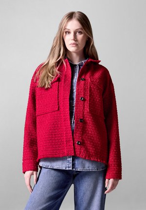 Rote strukturierte Jacke mit zwei großen Brusttaschen, schwarzen Knöpfen und einem Kragen, getragen über einem blauen Jeanshemd, kombiniert mit blauen Jeans.