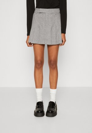 Kvinde iført en grå plissert mininederdel, hvide sokker, sorte chunky loafers og en sort langærmet top, stående på et gråt gulv mod en hvid væg.