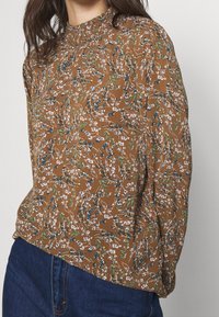 Brown floral blouse with a high neckline, featuring a pattern of white flowers and blue berries on a textured fabric. Long, billowy sleeves.