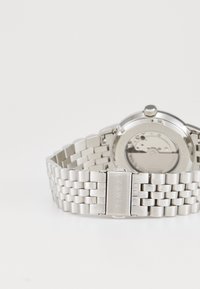 Montre en acier inoxydable argenté avec un bracelet maillé, boîtier rond, mouvement visible par l'arrière, et une boucle marquée "TIMEX."