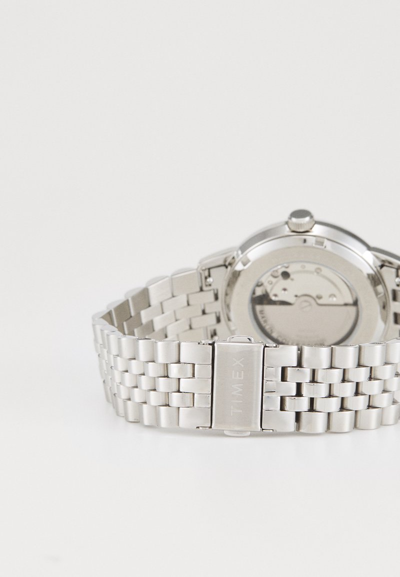 Silver stainless steel watch with a mesh bracelet, featuring a round case and an exposed movement display on the back.