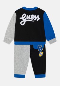 Set di abbigliamento da casa a due pezzi in nero, grigio e blu. Materiale morbido, con il logo "Guess" in pile bianco e una toppa sui pantaloni.