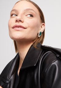 Pendientes rectangulares de plata con una gran piedra verde, usados con una chaqueta de cuero negra. Textura suave y diseño moderno.