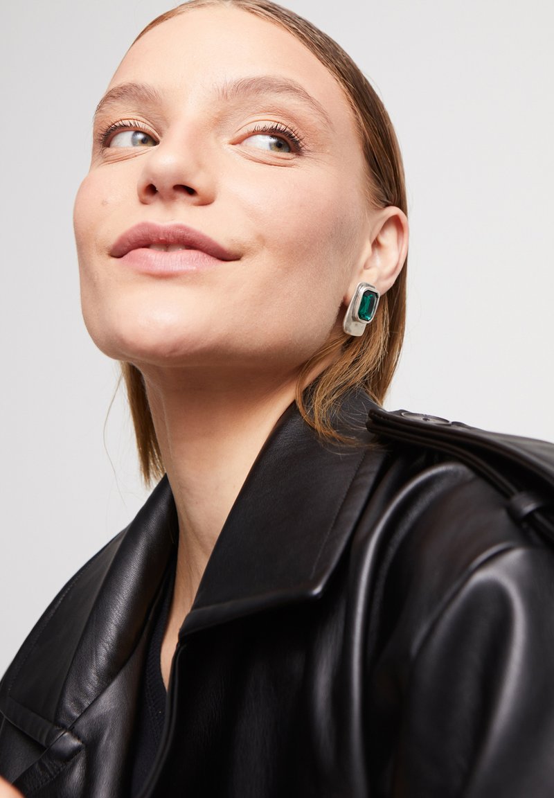 Pendientes rectangulares de plata con una gran piedra verde, usados con una chaqueta de cuero negra. Textura suave y diseño moderno.