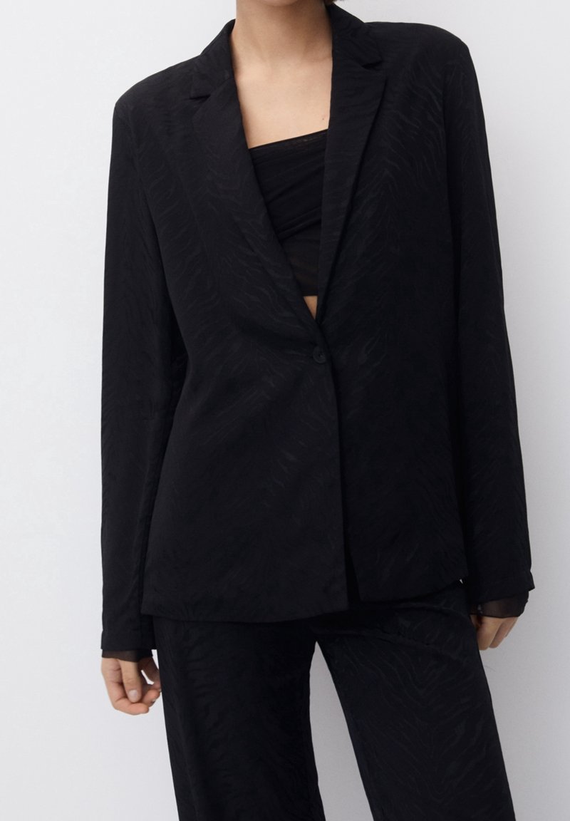 Femme portant un blazer noir texturé avec un bouton fermé et un pantalon assorti sur un fond uni clair.
