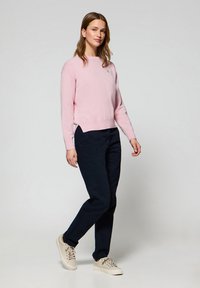 Ljusrosa tröja med rund halsringning, parat med mörkblå byxor. Modellen har på sig beiga sneakers och står framför en neutral bakgrund.