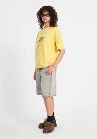 Femme portant des lunettes de soleil noires, un t-shirt graphique jaune, un short en jean gris au genou, des chaussettes noires et des chaussures à plateforme marron sur fond blanc.